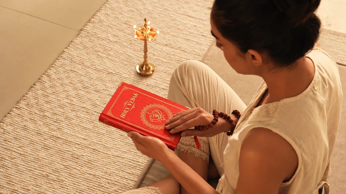 Art of Vedas: nainen istuu punotulla matolla ja lukee punakantista Holy Vedas -kirjaa messinkisen ayurvedisen öljylampun vieressä